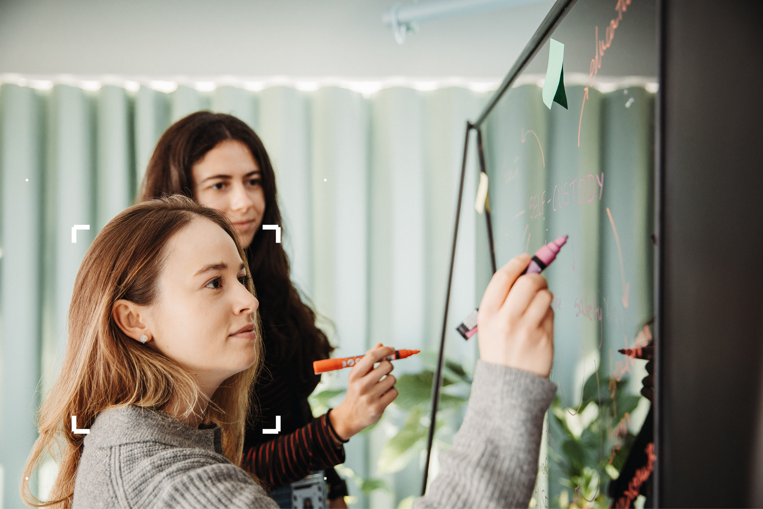 Two women writing on a white board with a green curtain in the background.