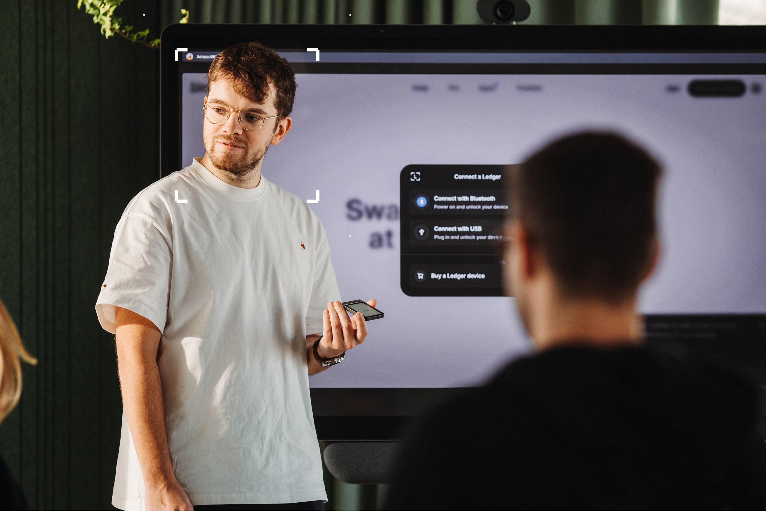 Man in white tshirt stood in front of a screen holding a Ledger signer device framed by a Ledger signer device