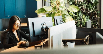 Woman sitting at a desk working, with a green wall behind her looking at 2 monitors